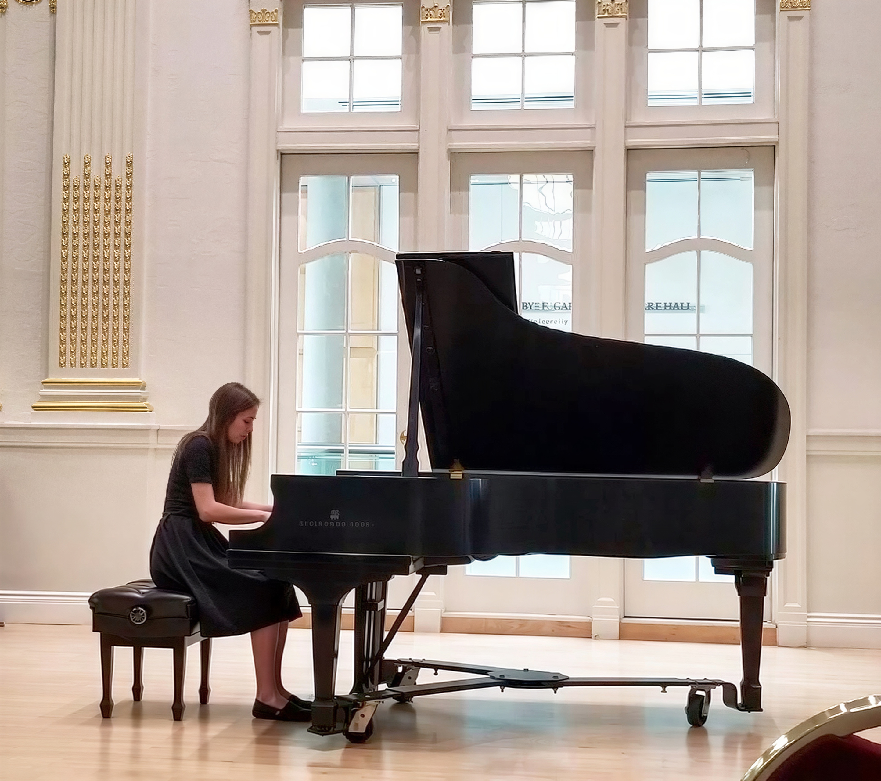 Kristianna Callahan performing on a Steinway grand piano in an ornate concert hall with tall windows and gold-trimmed walls